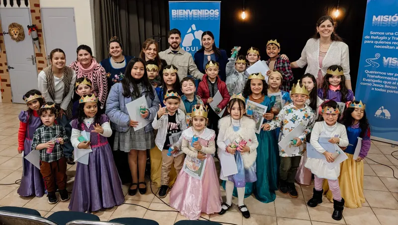Niños de Adulam disfrazados con coronas junto a sus maestras, frente al cartel de bienvenida de la iglesia